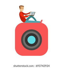 Young smiling man sitting on a big mobile app symbol and using his laptop colorful character vector Illustration