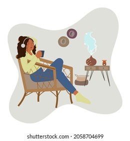 Young woman drinks tea and listening music on headphone sitting on armchair at cozy home while enjoying the aromatherapy from oil diffuser with flower smell. Aromatherapy Concept. Music therapy