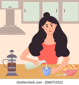 Young woman is having breakfast in the kitchen. Girl is sittting at the table and pouring milk into a mug of tea. There are porridge with strawberries and teapot on the table.