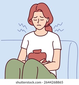 Jovem segura almofada de aquecimento contra o estômago enquanto senta enrolada no sofá. Ela sofre de cólicas menstruais e sintomas de período doloroso
