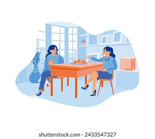 A young woman with long hair browsing her smart phone while having breakfast at home with her friend. Two women enjoying food while relaxing. Smiling woman friends drinking tea at home. 