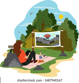 A young woman is relaxing in a park, drinking wine and watching a movie on the street. Street cinema