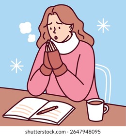 Young woman sits at table reading a book and warming hands near hot drink in cold unheated room