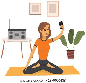 Young woman sitting in lotus position with smartphone her in hands. Yogi is posing for self portrait. Girl doing yoga and meditating during photo shoot. Lady is photographed on phone camera