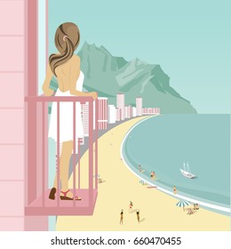 Young woman stands on balcony of hotel looking at ocean coast with people on beach, mountains and buildings along road