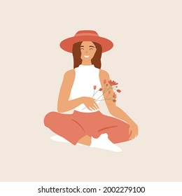 Young woman in summer hat sitting and holding flowers branch in hand. Beautiful happy girl in stylish clothes and shoes. Character for farming, gardening, countryside concepts