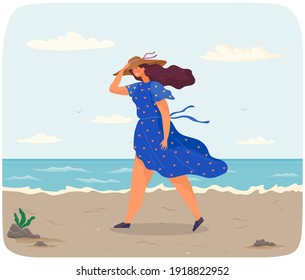 Young woman walking alone on beach. Lady hold her hat on head because of windy weather. Person dressed in blue dotted dress. Beautiful landscape of ocean bank. Girl is walking during strong wind