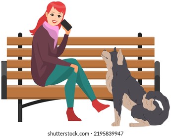 Young woman walks with dog in city park. Concept of outdoor walks during cold season. Pet is best friend, animals from shelter. Doggy owner sitting on bench on street outdoors and talking on phone