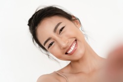 Closeup portrait of beautiful young Asian smiling with white teeth and taking selfie of herself over white wall background. Natural Beauty and Healthy Woman.