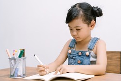 Portrait of cute little Asian girl writing or drawing, coloring pages, in notebook at desk on white background. School kid girl to do homework. Elementary school and homeschooling concept.