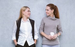 Cheerful young two women talking together during coffee break standing in modern office. Caucasian girl communicates with workmate hold coffee cups enjoy conversation, discuss work or personal.