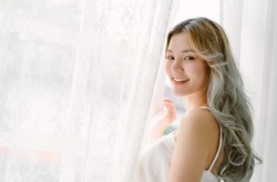 Portrait of beautiful Asian girl hair coloring in blondE standing near a window white curtains and turning to look and smile at camera. Alluring japanese woman posing near curtain in bedroom at home.