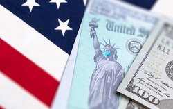 United States IRS Stimuls Check with Statue of Liberty Wearing Medical Face Mask Resting on American Flag.