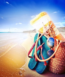 Summer accessories, swimsuit, sun glasses, bag and flip-flops. Closeup of summer beach bag and straw hat on sandy beach.