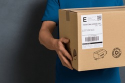 Closeup hands of delivery man holding package to deliver. Courier hand holding brown box isolated on grey background. Detail of delivery man carrying cardboard parcel with label with copy space.