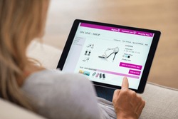 Closeup of woman doing online shopping on digital tablet at home. Rear view of woman hand touching screen while selecting shoes on ecommerce portal. Lady use e-commerce webshop to buy shoes.