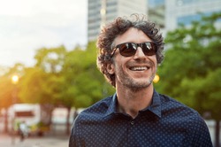 Portrait of smiling man wearing sunglasses and looking away in the city streets. Cheerful mature businessman walking with a big smile on face. Happy man in blue shirt and glasses enjoying the sunset.