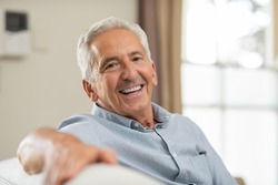 Portrait of happy senior man smiling at home. Old man relaxing on sofa and looking at camera. Portrait of elderly man enjoying retirement.