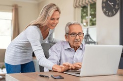 Senior man and mature daughter smiling and looking at laptop at home. Happy daughter teaching and showing new computer technology to her old dfather. Old man learn to use computer.