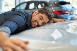 Happy young man hugging his new car in showroom. Satisfied guy with closed eyes embracing the hood of the automobile. Dreaming man lying on car bonnet hugging it.