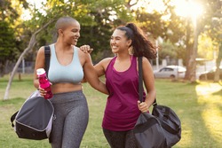 Cheerful smiling friends in sportswear holding gym bag and bottle in park. Multiethnic women going to park for fitness workout. Two curvy girls walking after exercise session outdoor at sunset.