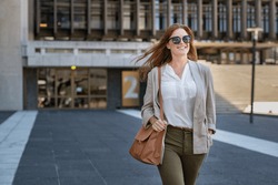 Portrait of successful happy woman on her way to work on street. Confident business woman wearing blazer carrying side bag walking with a smile. Smiling woman wearing sunglasses and walking on street.