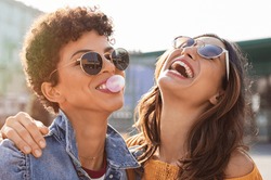 Young latin woman laughing while friend inflating bubble gum. Closeup face of multiethnic friends enjoying outdoor street. Brazilian girl laughing and blowing chewing gum with friend embracing her.