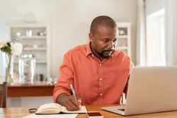 Mature man working on laptop while taking notes. Businessman working at home with computer while writing on agenda. African man managing home finance, reviewing bank account and using laptop.