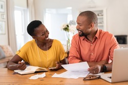 Mature couple sitting and managing expenses at home. Happy african man and woman paying bills and managing budget. Black smiling couple checking accountancy and bills while looking at each other.