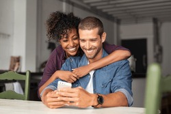 Smiling young couple embracing while looking at smartphone. Multiethnic couple sharing social media on smart phone. Smiling african girl embracing from behind her happy boyfriend while using cellphone