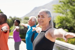 Portrait of happy senior woman practicing yoga outdoor with fitness class. Beautiful mature woman stretching her arms and looking at camera. Portrait of smiling lady with outstretched arms at park.