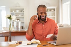 Smiling african man using laptop at home while checking home finance. Happy mature man looking at invoice while talking on phone with bank. Man checking receipt and bill while talking at phone.