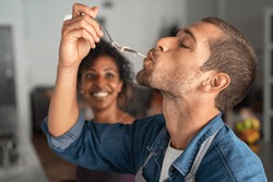 Man tasting spaghetti pasta while smiling woman look at him. Closeup face of young man tasting meal while cooking at home. Handsome guy eating noodles with fork in kitchen and feel his recipe.