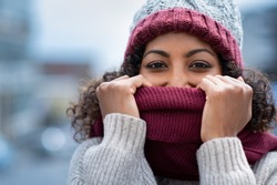 Closeup face of happy african girl holding woolen scarf with hands over nose to protect from the frost. Beautiful young woman in warm winter knitted clothes covering her face and looking at camera.