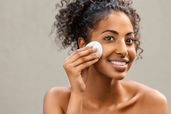 Young pretty african american woman taking off makeup with cotton wipe sponge. Smiling girl cleaning face isolated over background. Black young woman cleansing face, daily healthy beauty routine.