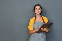 Happy smiling waitress ready to take customer order while looking at camera isolated on grey wall. Mature woman wearing apron while writing on clipboard standing against gray background and copy space