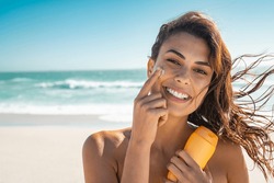 Beautiful young woman at beach applying sunscreen on face and looking at camera. Beauty latin girl applying suntan lotion at sea. Portrait of happy woman with healthy skin applying sunblock on cheek.
