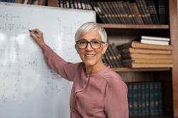 Portrait of happy mature professor teaching mathematics to students in a library. Senior smiling woman solving math problem while writing on white board. Portrait of tutor looking at camera.