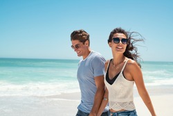 Beautiful woman with man wearing sunglasses walking on beach.. Young couple enjoying honeymoon after marriage at sea. Happy casual couple holding hands and walking at the beach with copy space.