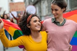 Young women on street enjoying holding gay pride flag during protest. Smiling multiethnic women enjoying during march on street for lgbt rights. Diversity, tolerance and gender identity concept.