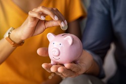 Close up of man holding pink piggybank while woman putting coin in it. Indian young couple saving money for their wedding. Close up of woman hand putting euro money in piggy bank to save.