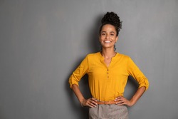 Portrait of young african woman standing with hands on waist and looking at camera. Confident stylish latin girl smiling isolated against grey background. Beautiful woman with copy space on gray wall.