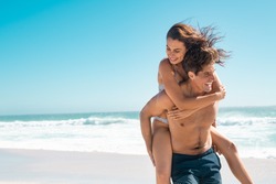 Young couple enjoying vacation at beach, copy space. Portrait of mid smiling man carrying girlfriend on his back along the sea shore. Boyfriend giving piggyback ride to happy woman having fun together