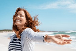 Happy mature woman with arms outstretched feeling the breeze at beach. Beautiful middle aged woman with arms up dancing on beach. Mid lady feeling good and enjoying freedom at sea, copy space.