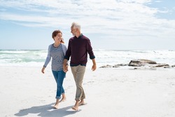 Senior couple walking and looking at each other at beach. Happy mature couple in love walking barefoot at sea shore. Loving retired man with beautiful woman relaxing at the ocean with copy space.