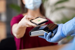 Cashier hand holding credit card reader machine and wearing disposable gloves while client holding phone for NFC payment. Woman wearing face mask while paying with smartphone during Covid-19 pandemic.