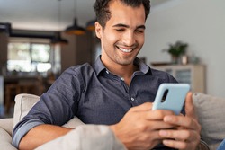 Cheerful businessman using smartphone while sitting on sofa at home. Handsome young indian man sitting on couch reading messages on mobile phone. Hispanic guy working from home with cellphone.