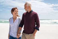 Happy senior couple walking on the beach in a sunny day. Smiling mature couple looking at each other on beach during sunset with copy space. Retired man in love with his wife relaxing during vacation.