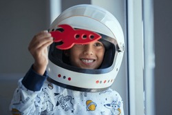 Happy smiling boy wearing an astronaut helmet costume and playing with a spaceship. Child playing with toy rocket near the window. Kid playing with red toy spaceship and dreaming outer space.