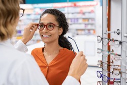 African young woman in optic store choosing new cool-down glasses with optician. Mixed race girl trying new eyeglasses with the help of the pharmacist. Multiethnic woman trying rest spectacle frame.